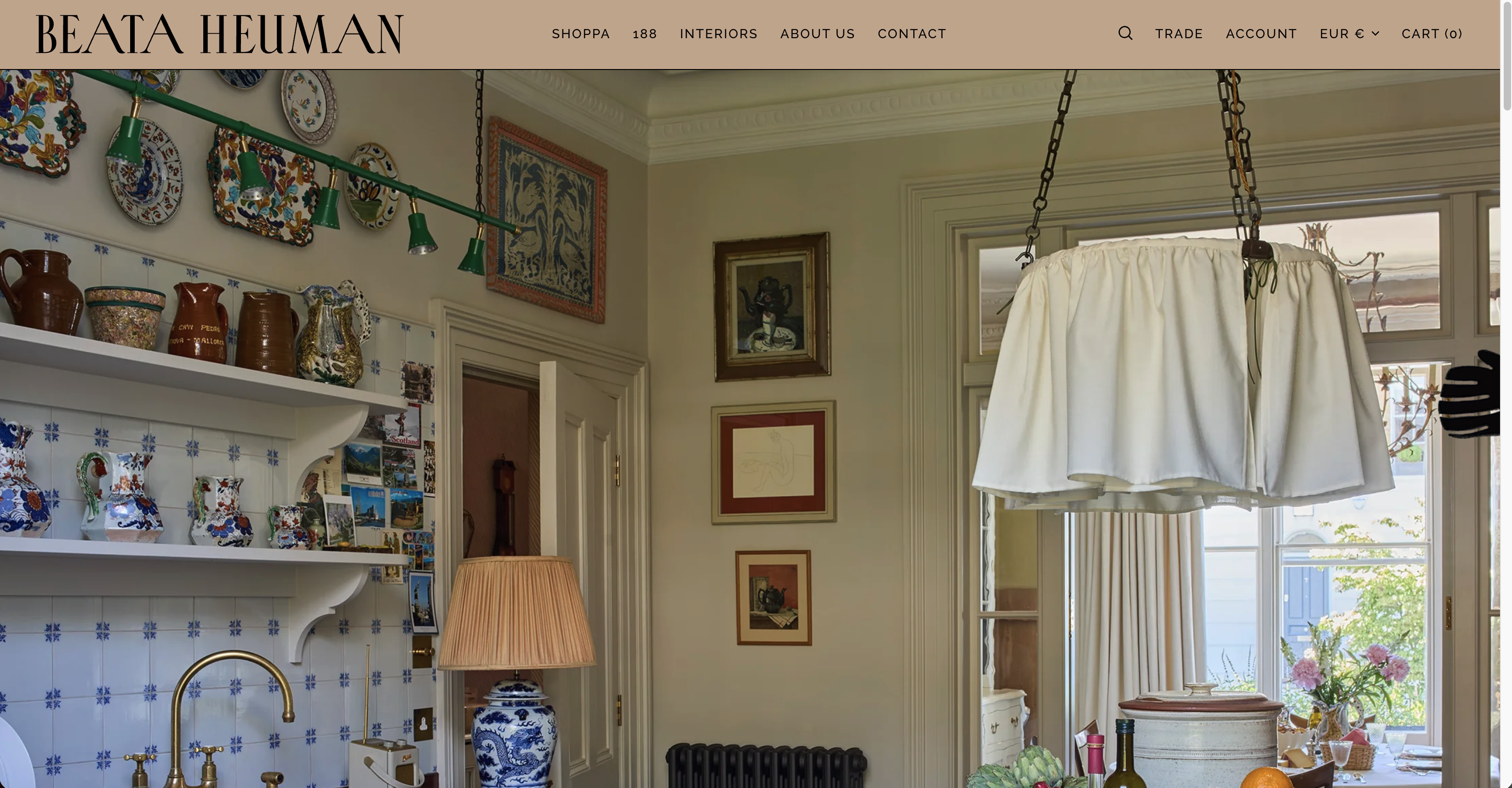 Beata Heuman homepage, with a full-width photo of a kitchen with patterned plates, green wall lights and a ruffled pendant lamp under a tan navigation bar